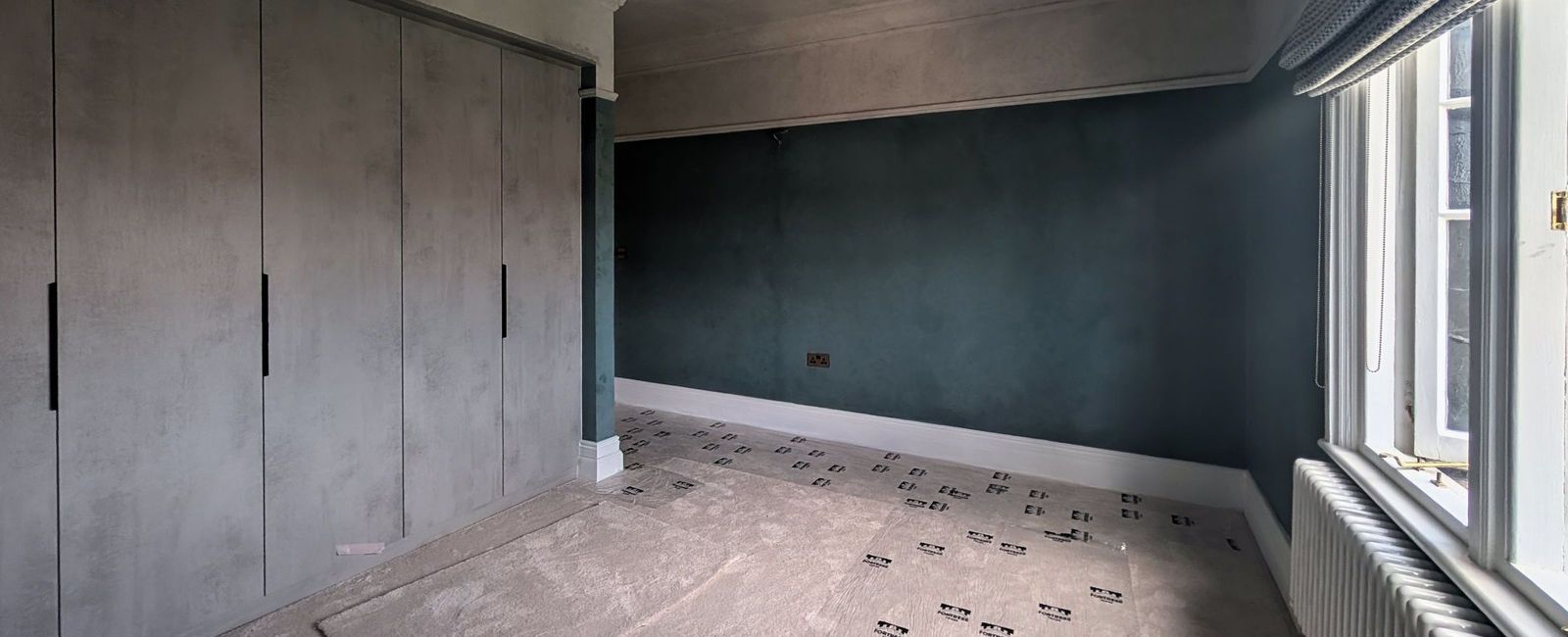 A run of full height fitted wardrobe doors in pale grey green along one wall, a deep teal wall to the right and a sash window